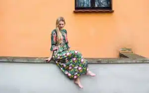 Iris Bergkrantz sitter på en mur, tittar nyfiken ner i marken. Bakgrunden är en stor orange cementvägg. hon har en blommig klänning på sig.