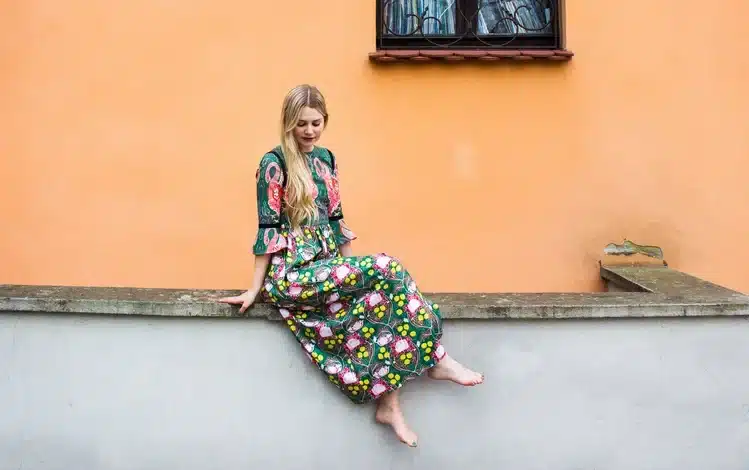 Iris Bergkrantz sitter på en mur, tittar nyfiken ner i marken. Bakgrunden är en stor orange cementvägg. hon har en blommig klänning på sig.